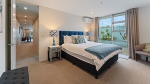 A bedroom with a large bed, a nightstand, a bench, and a view of the outdoors. - Waimahana 5 - Taupo Holiday Apartment (Taupo)