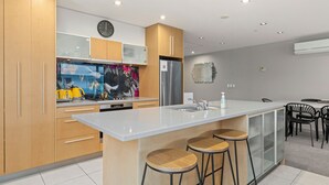 A modern kitchen with wooden cabinets, a stainless steel refrigerator, a large island with stools, and a dining area with chairs. - Waimahana 5 - Taupo Holiday Apartment (Taupo)
