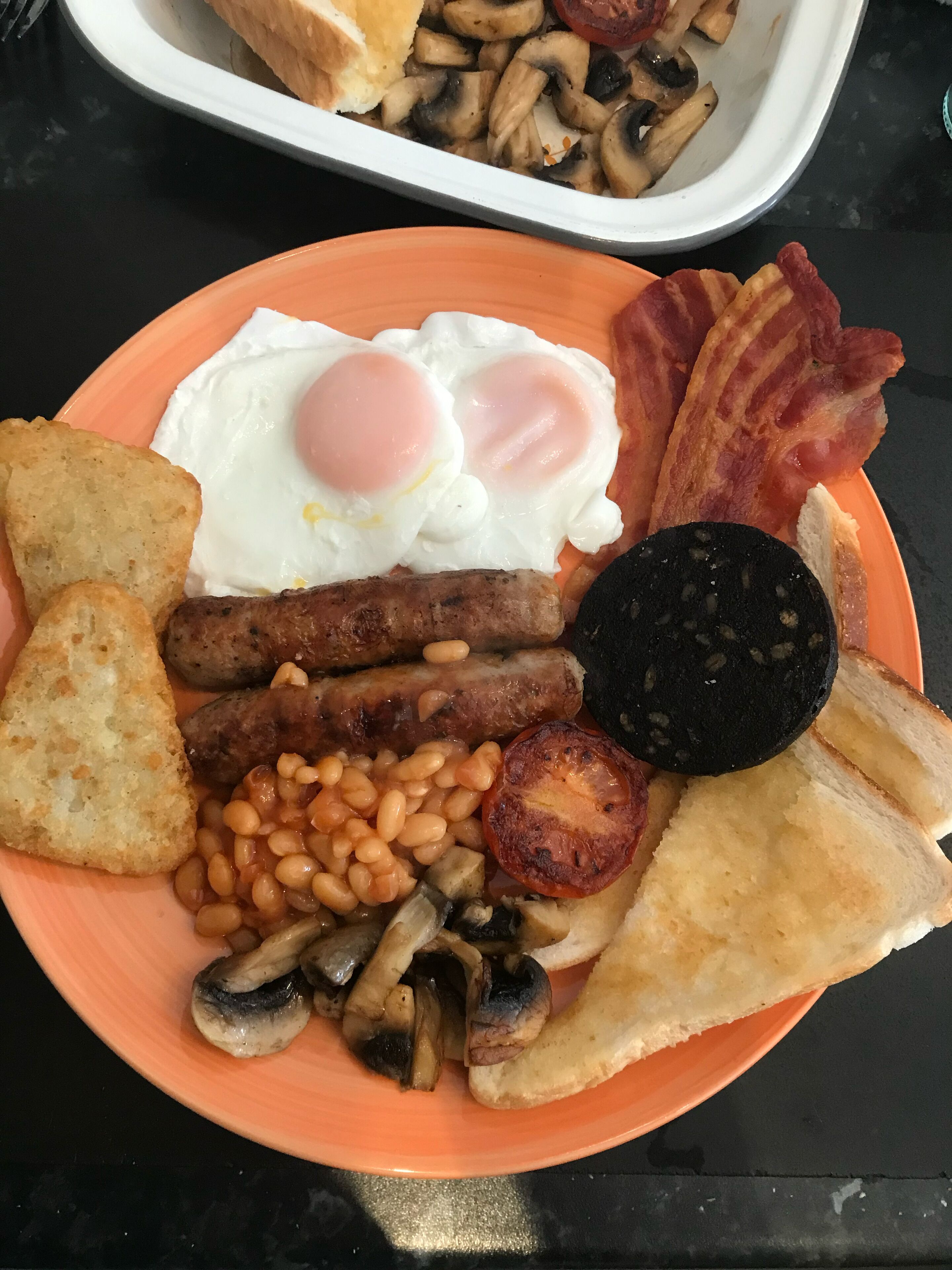 Un plato con huevos, tocino, salchichas, frijoles, champiñones, tostadas y una rebanada de morcilla.