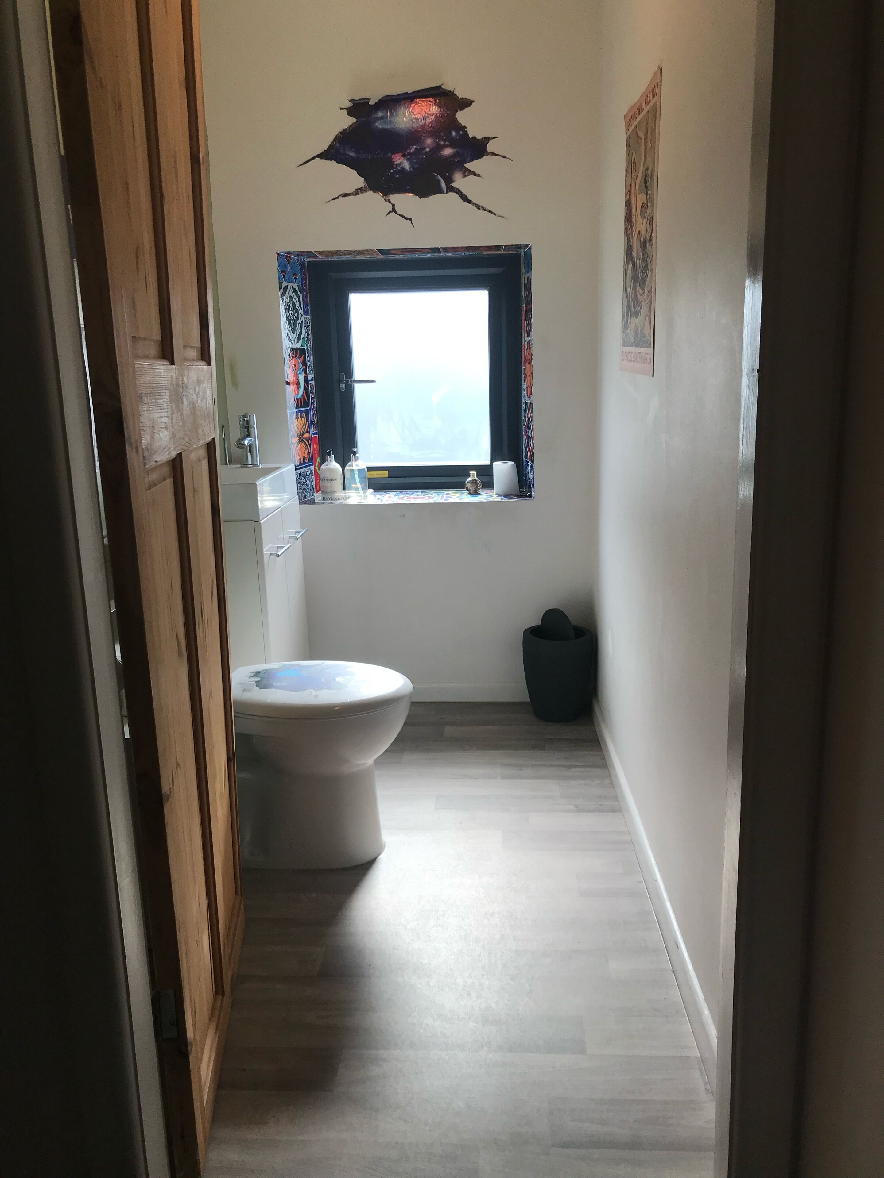 A narrow hallway leading to a bathroom with a toilet, a window with curtains, and a wall decal.