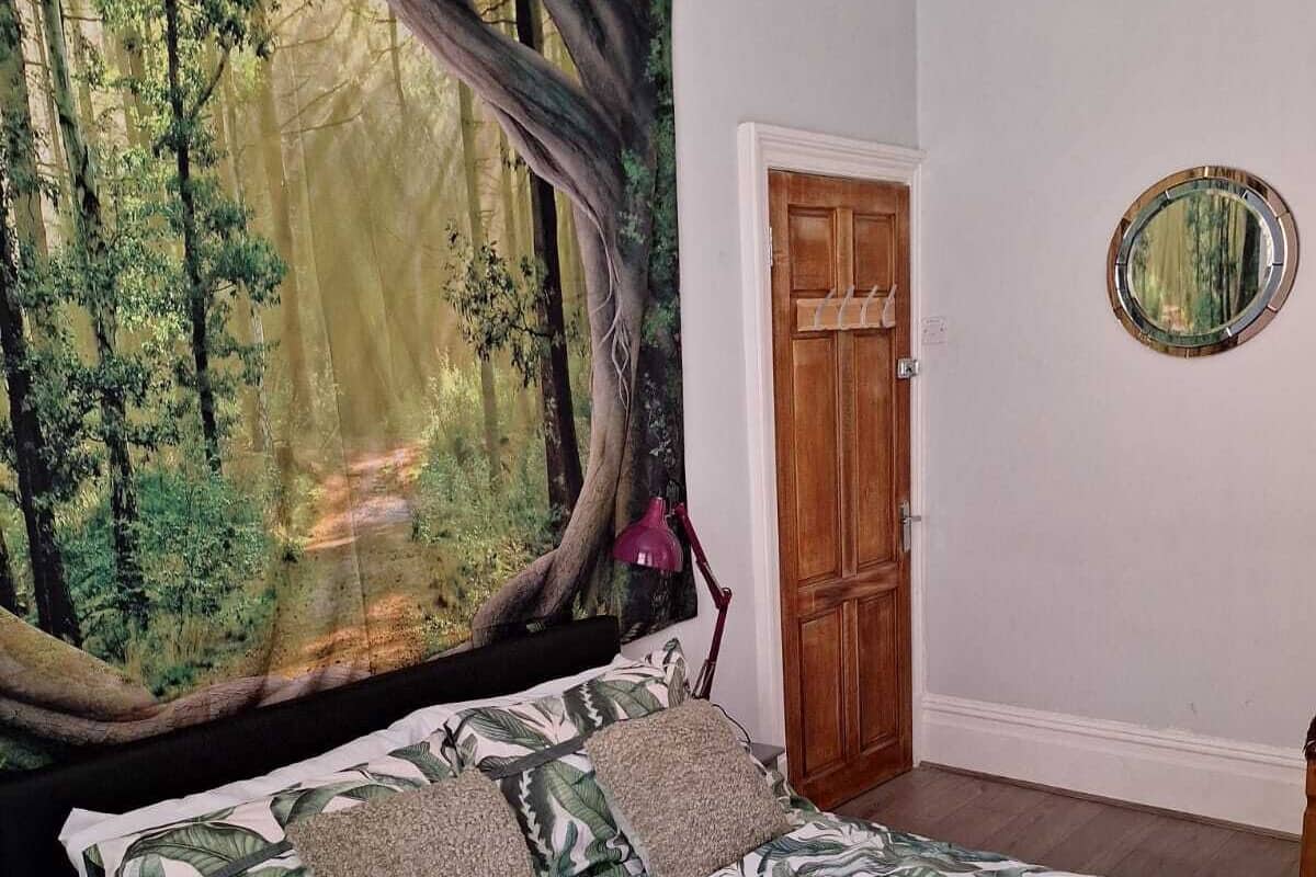 Une chambre à coucher avec un lit orné d’un motif de feuilles tropicales, une grande tapisserie sur le thème de la forêt, une porte en bois et un miroir rond accroché au mur.