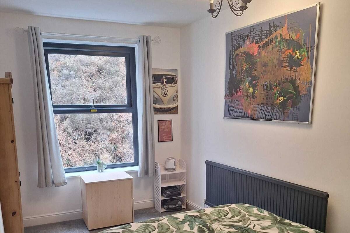 Une chambre avec une grande fenêtre, un radiateur, un lit avec une couette à motifs végétaux, une étagère avec divers objets et un tableau mural.