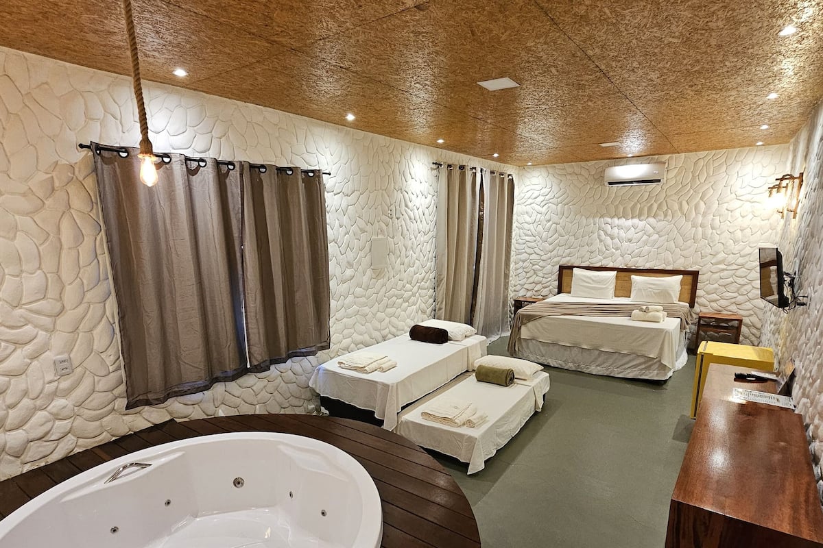 A room with a large bathtub, two beds, a wooden vanity, and stone-textured walls.
