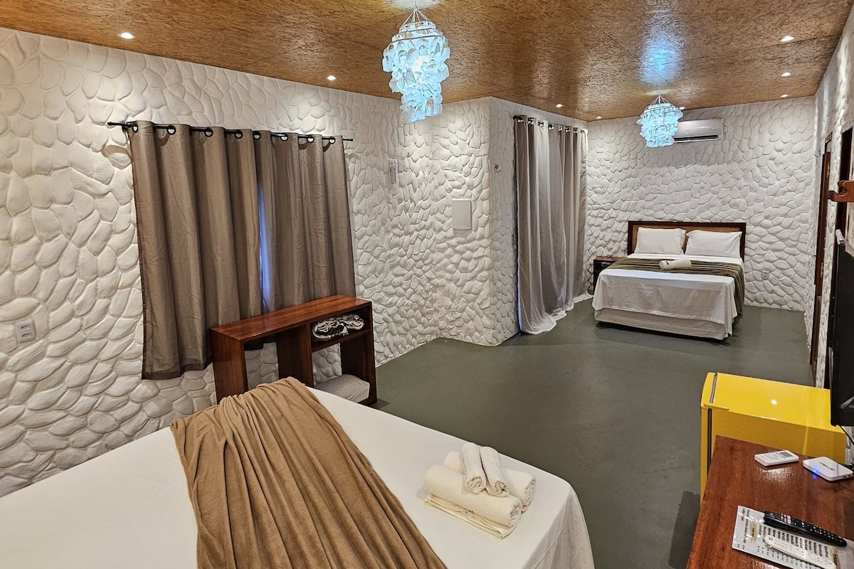 A hotel room with a wooden ceiling, stone-textured walls, a bed with a brown blanket, a bedside table with a remote control, and a desk with a computer.