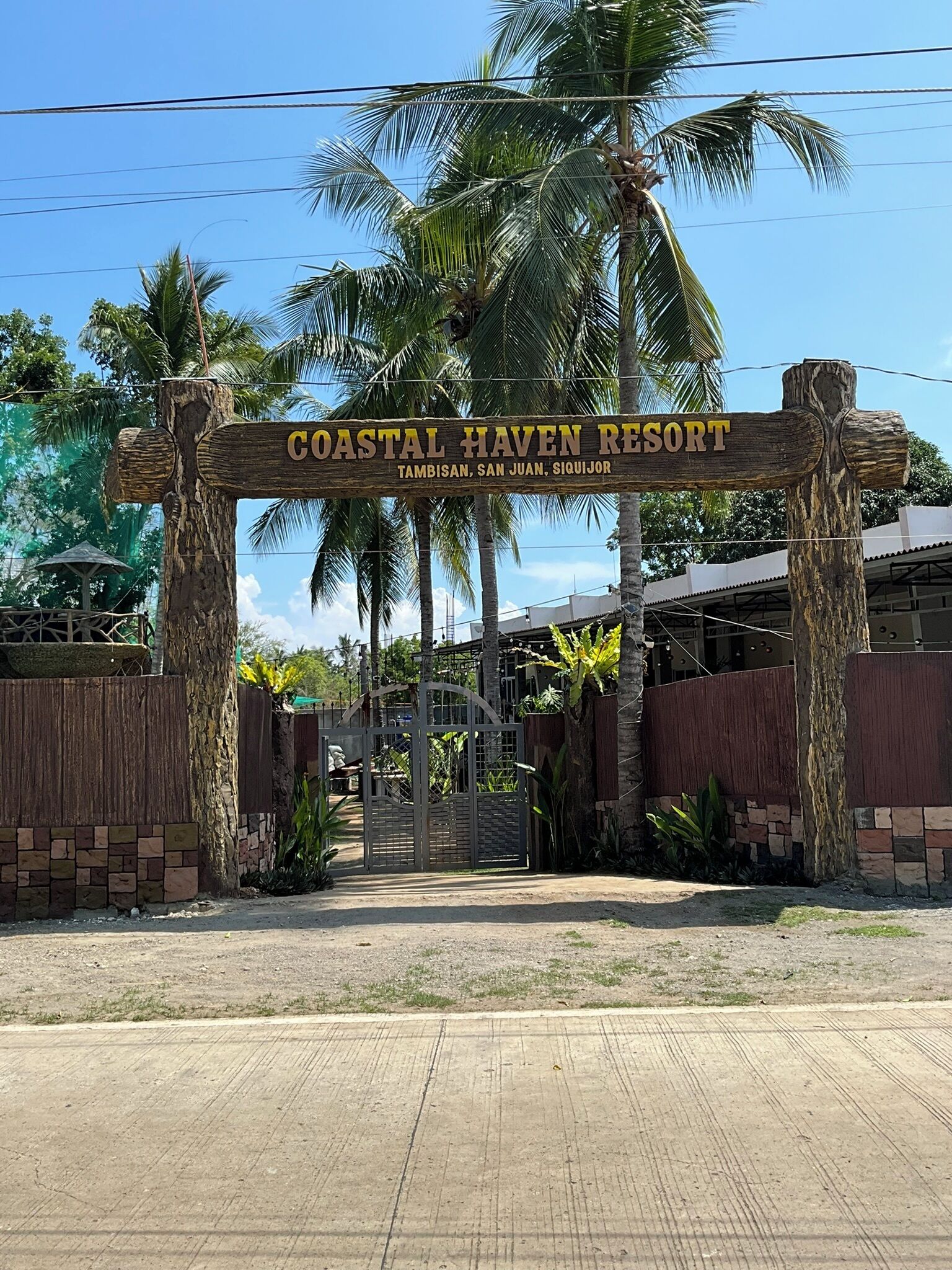 Un letrero de madera con la inscripción 'Coastal Haven Resort', rodeado de palmeras y una entrada con puerta de acceso.