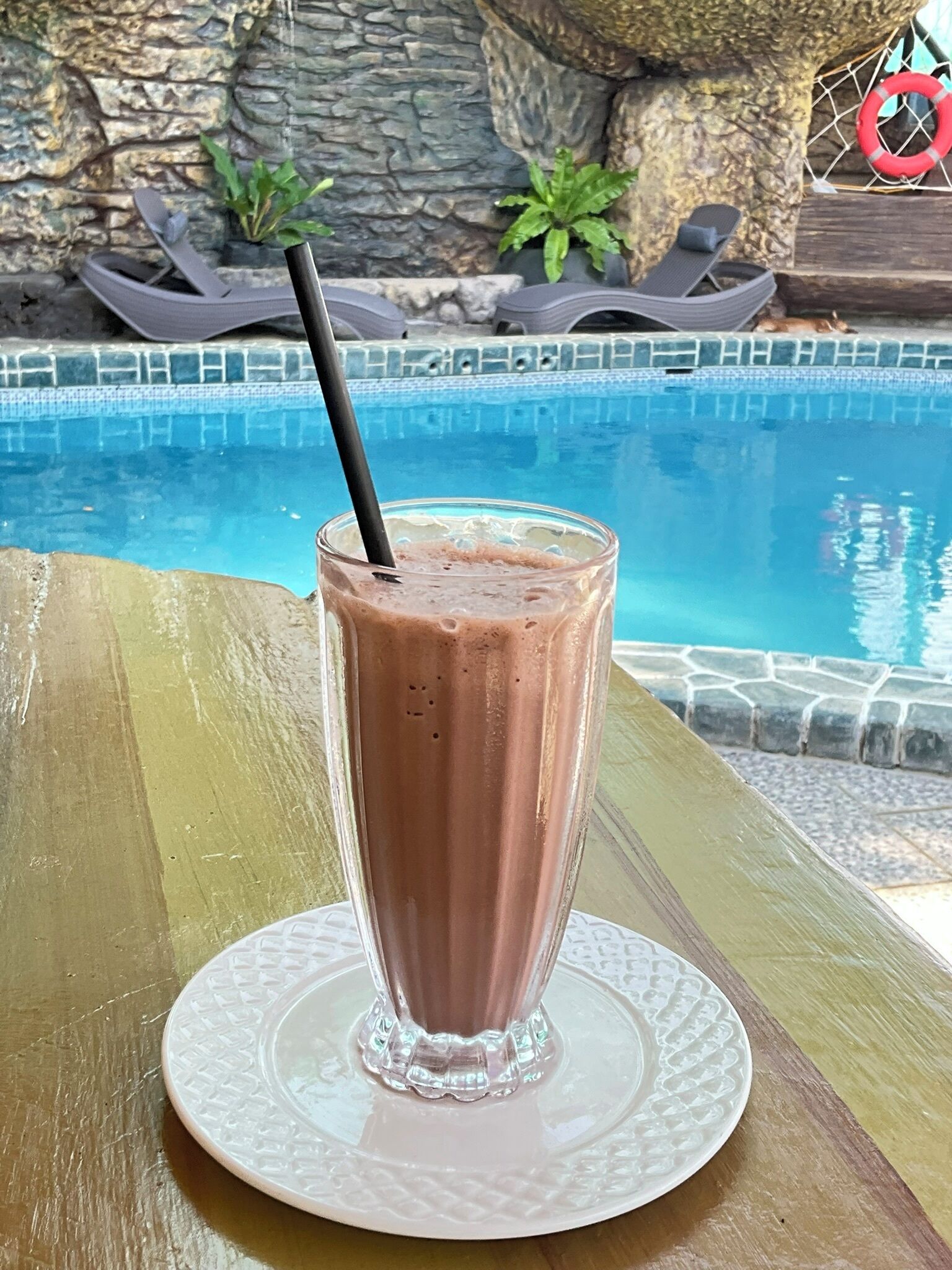 A glass of chocolate milkshake with a straw on a white plate by a poolside.
