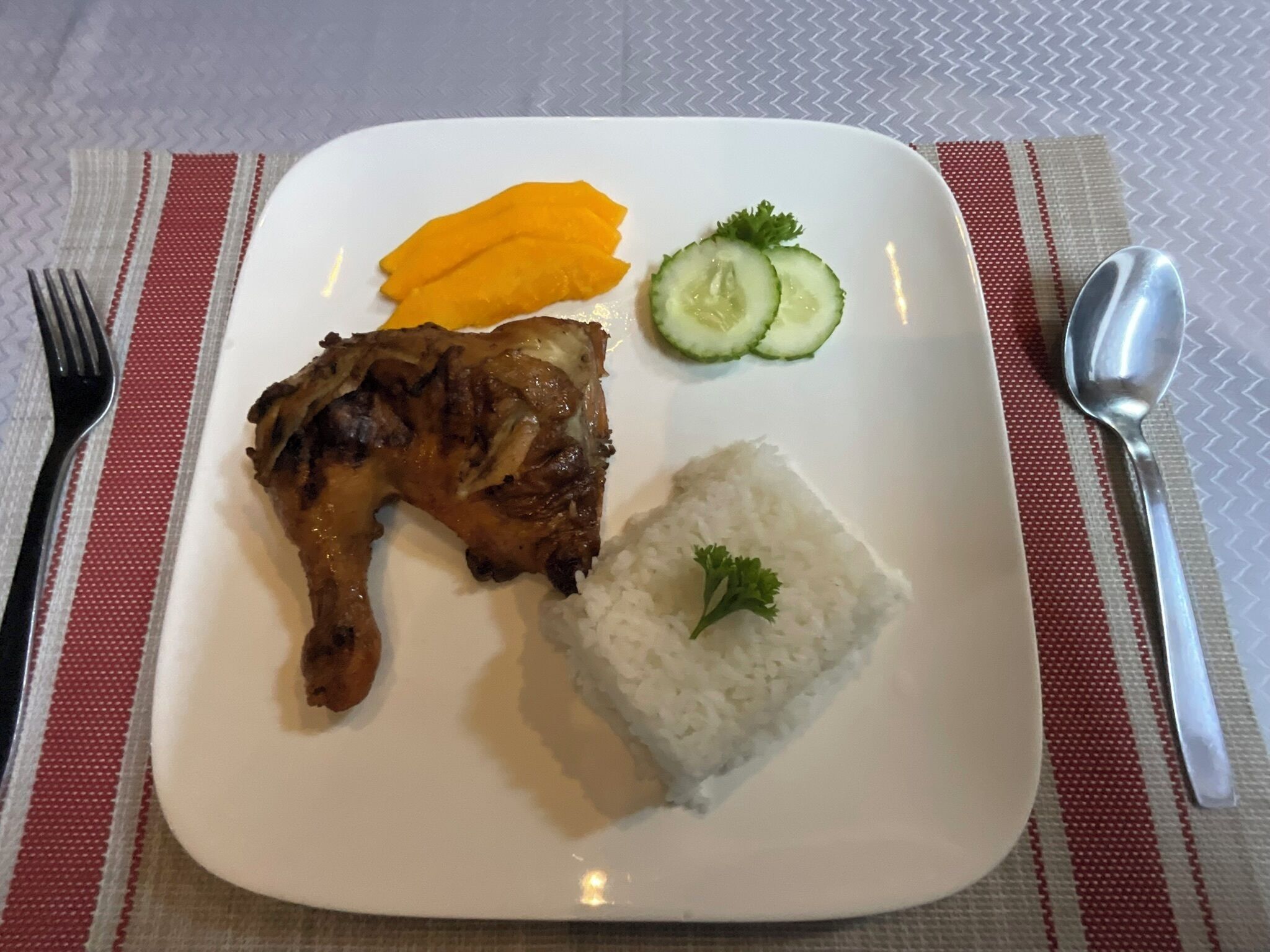 A plate with grilled meat, white rice, mango slices, and cucumber slices.