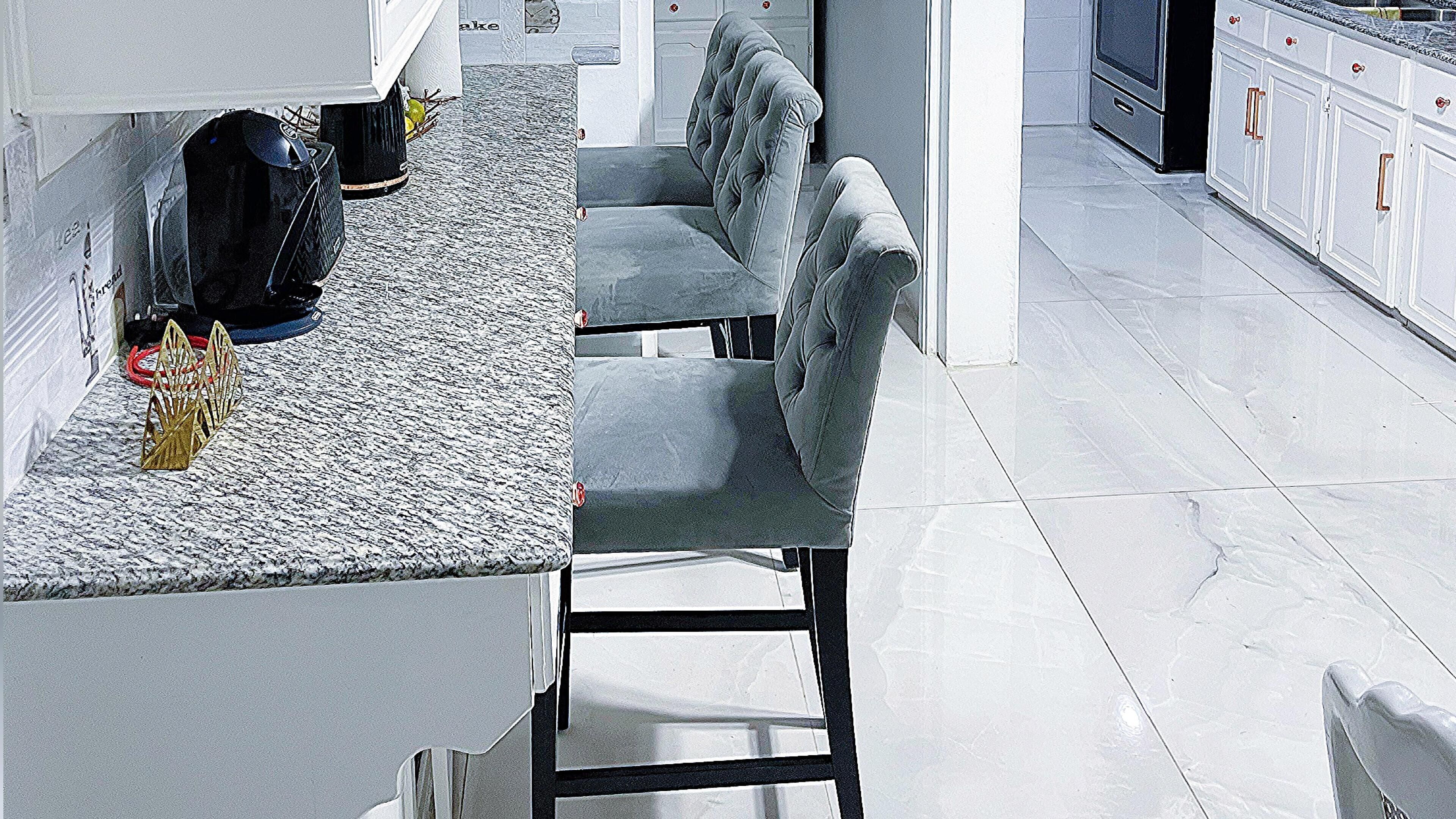 A modern kitchen with a granite countertop, two grey upholstered chairs, and a white cabinet with red handles.