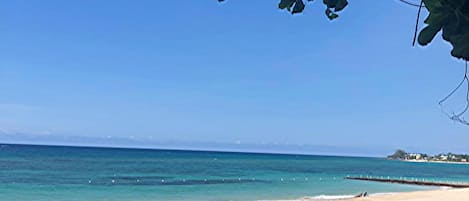 A beach with clear blue water, a sandy shore, and a distant shoreline with buildings.