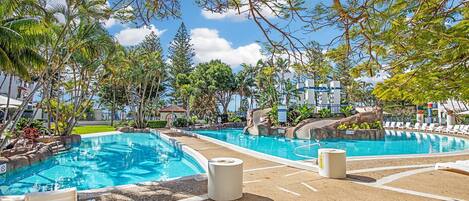 Un espace piscine comprenant plusieurs piscines, des chaises longues et un jardin aux allures tropicales.