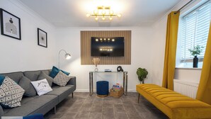 A living room with a grey sofa, a blue ottoman, a yellow bench, a glass coffee table, a TV mounted on a wooden panel, and a window with yellow curtains. - Comfy House - Sleeps 14 - 14 min to City Centre (Manchester)