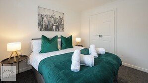A neatly made bed with green bedding, two white lamps on side tables, and a cityscape painting on the wall. - Comfy House - Sleeps 14 - 14 min to City Centre (Manchester)