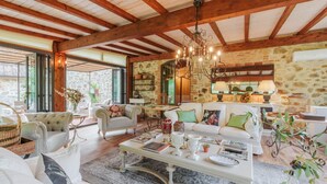Ein geräumiges Wohnzimmer mit hoher Decke, freiliegenden Holzbalken und einer Steinwand. Es gibt mehrere Sitzbereiche mit weißer und grüner Polstermöbel.
