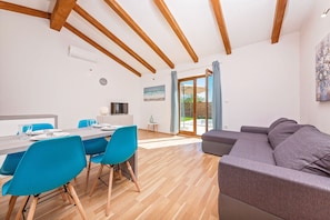 Ein modernes Wohnzimmer mit Holzbalkendecke, einem grauen Sofa und einem Essbereich mit blauen Stühlen und einem Holztisch.