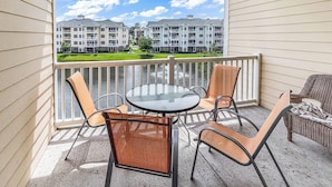 A balcony with a round glass table and four chairs, overlooking a building with multiple balconies and a grassy area. - Myrtlewood Top floor Condo 2BR • 4 Beds • 2 full Baths • with Elevator (Myrtle Beach)
