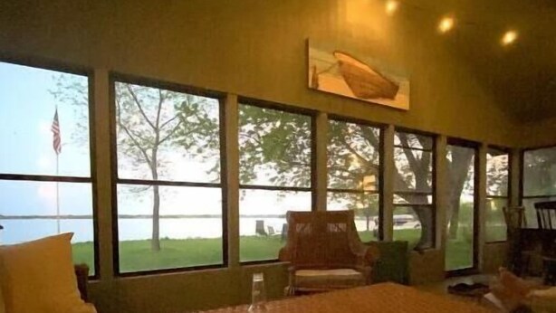 A cozy living room with a large window overlooking a grassy area and a flag. There is a wooden table with a bottle and papers, a sofa, and a chair. - Escape to this peaceful lakefront getaway on Lake Hendricks (White)