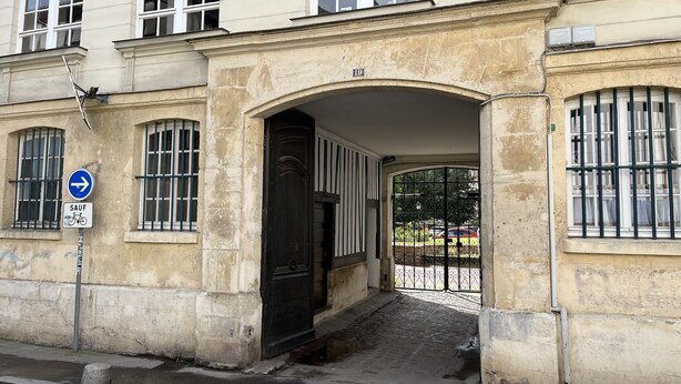 Une entrée d’immeuble avec un arc en pierre, un panneau indiquant une direction, et une zone délimitée par une grille, avec une voiture visible au fond.