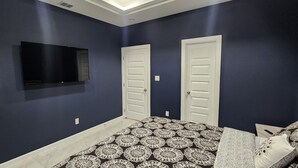 Un dormitorio con una colcha estampada, dos puertas blancas, un televisor montado y una lámpara en el techo.