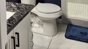 Un baño con un inodoro blanco, un armario blanco, una ducha con puerta de cristal y un suelo de baldosas de mármol.