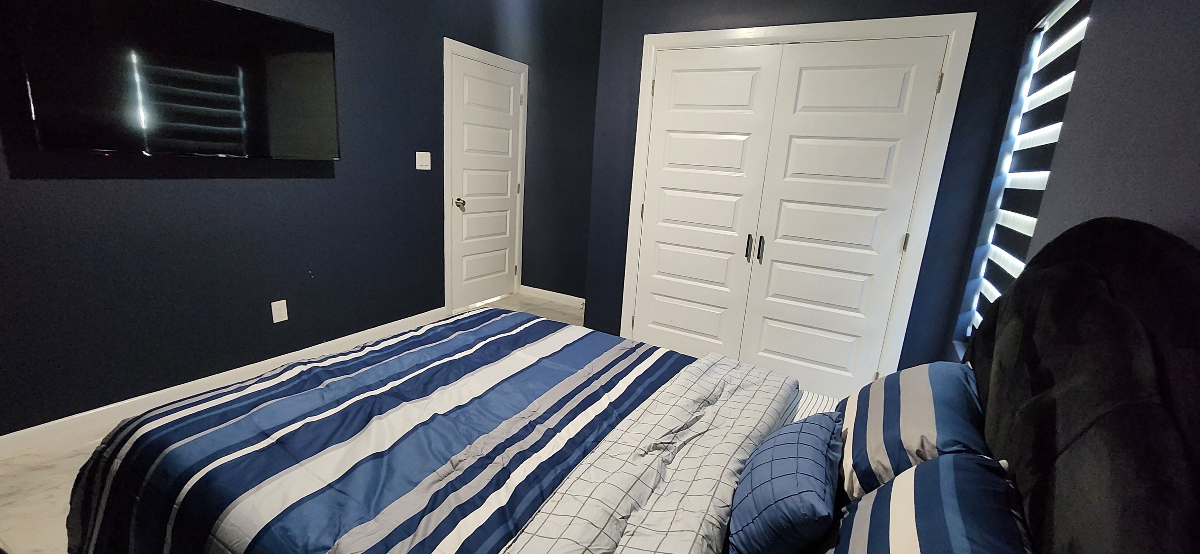 A bedroom with a bed, two white doors, and a window with blinds.