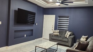 Un salón con paredes azul oscuro, un televisor de pantalla plana montado en la pared, un sofá gris con cojines estampados, una mesa de centro y un ventilador de techo.