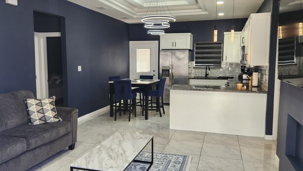 Una sala de estar moderna con un sofá gris, un comedor con sillas, una cocina con gabinetes blancos y un piso de baldosas de mármol.