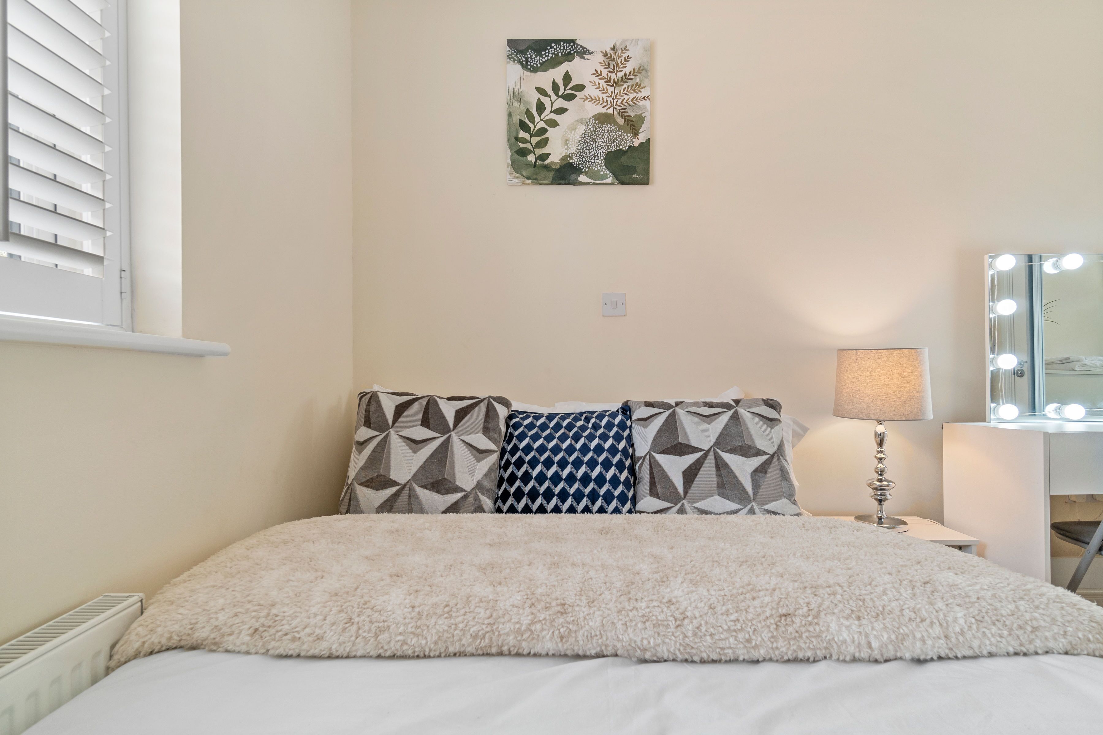 A neatly arranged bedroom with a bed, pillows, a bedside lamp, and a wall-mounted mirror with lights.