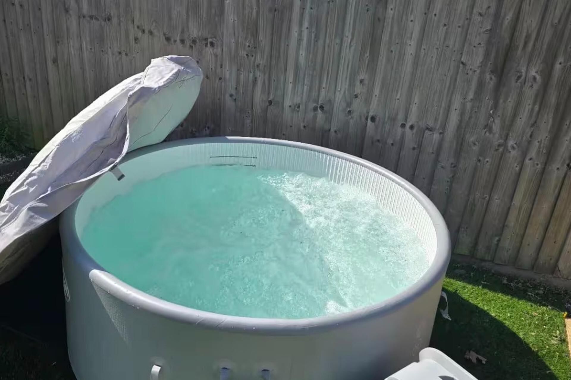 A large, light-colored hot tub with a partially open lid, surrounded by a wooden fence.