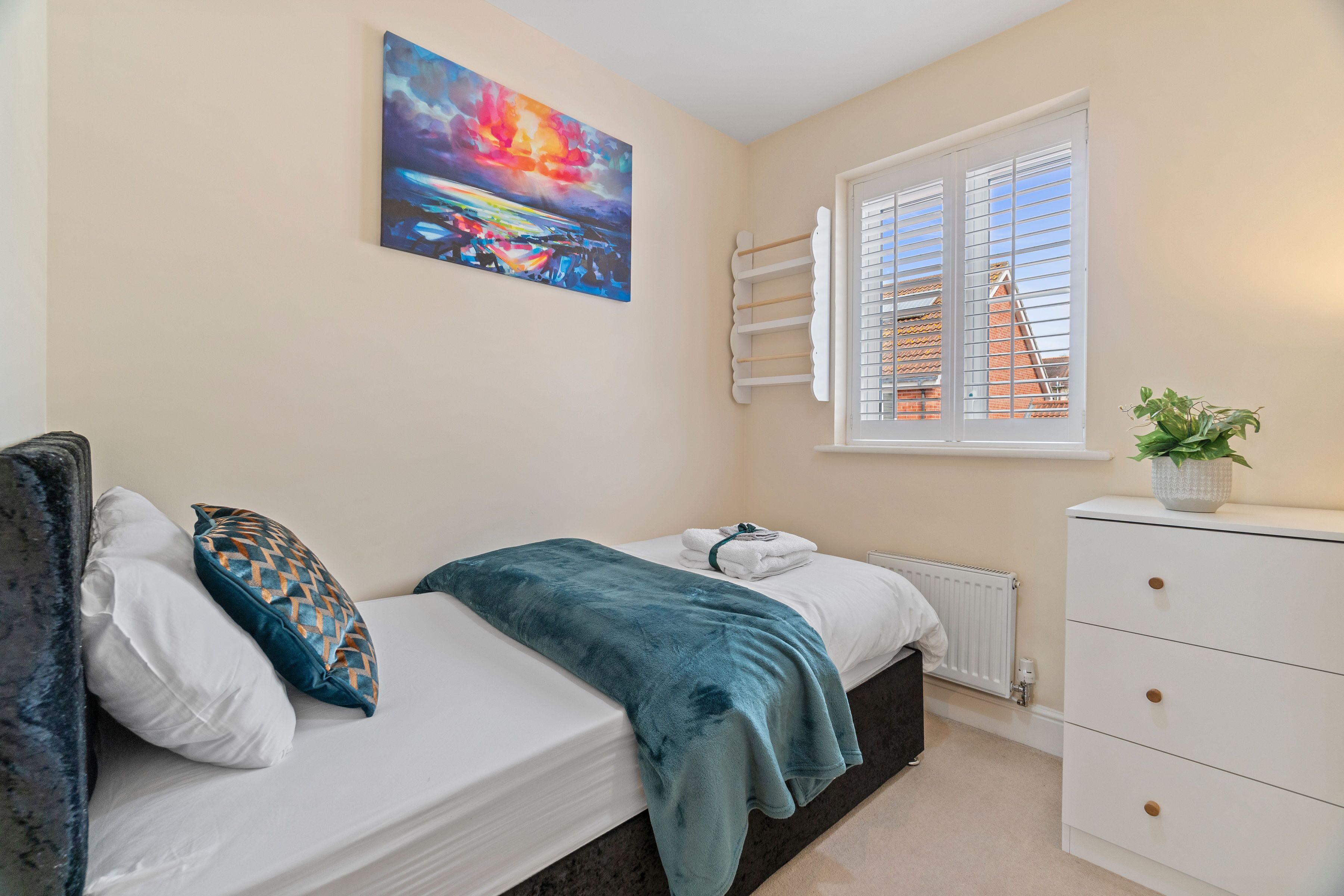 A bedroom with a bed, a window with blinds, a radiator, a dresser, and a painting on the wall.