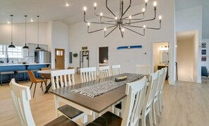 A modern kitchen with white cabinets, a central island, pendant lights, and a dining area with a large table and white chairs.