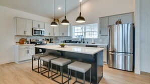 A modern kitchen with white cabinets, stainless steel appliances, a large island with bar stools, pendant lighting, and a view to the outside. - Branson Vacation Rental w/ Table Rock Lake Views!  (Branson)