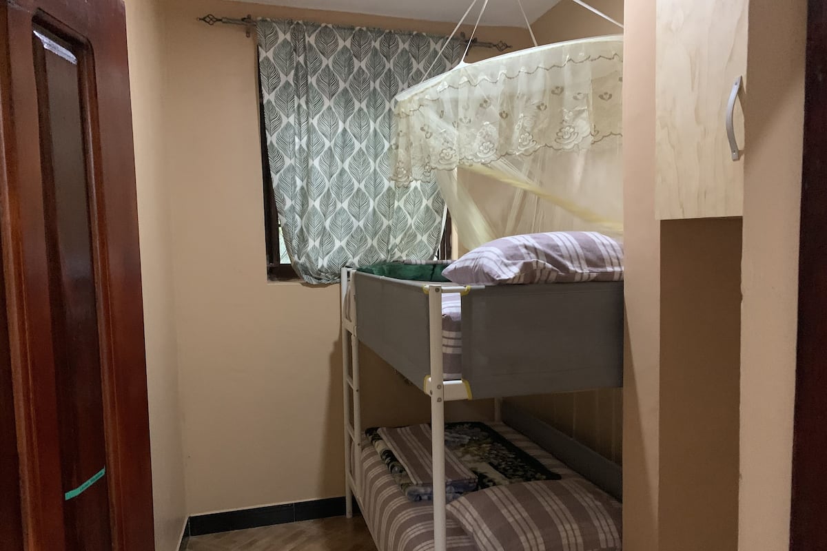 A small, compact room with bunk beds, a window with patterned curtains, and a door.