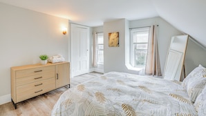 A bedroom with a bed, a dresser, a wall-mounted light, a window with curtains, and a painting on the wall. - Weymouth Apartment: Close to Beaches & Boston! (Weymouth)
