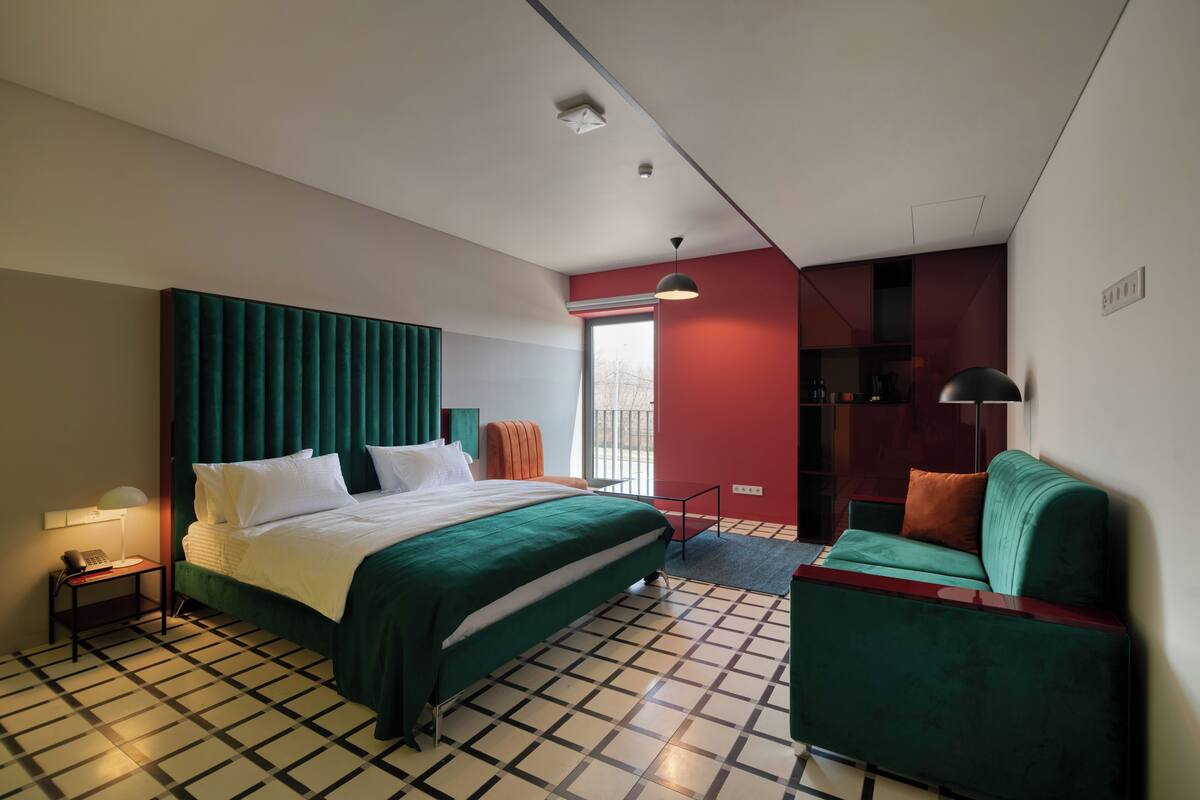 Ein Hotelzimmer mit einem großen Bett, einem grünen Sessel, einem kleinen Tisch mit Lampe und einer roten Akzentwand.