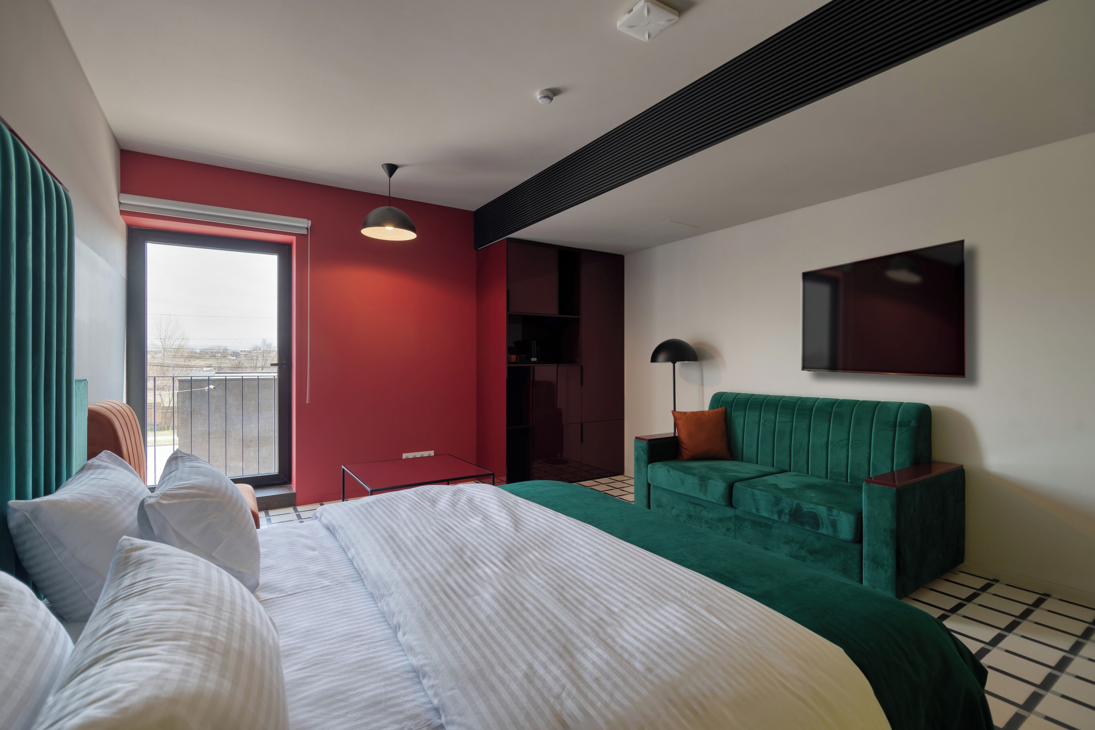 Una habitación de hotel moderna con una cama grande, un sofá verde, un televisor montado en la pared y una ventana con vistas a la ciudad.
