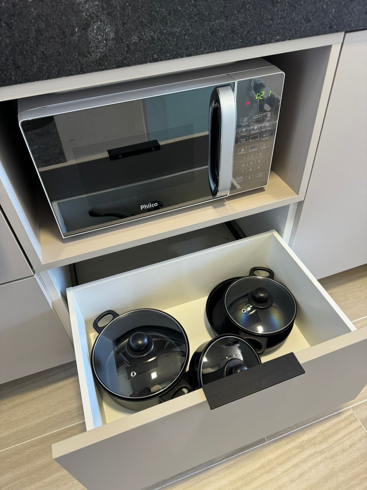A kitchen with a microwave and a drawer containing three black pots.