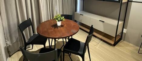 A room with a round wooden table, four black chairs, a potted plant, a built-in bench with storage, and a large window with blinds.