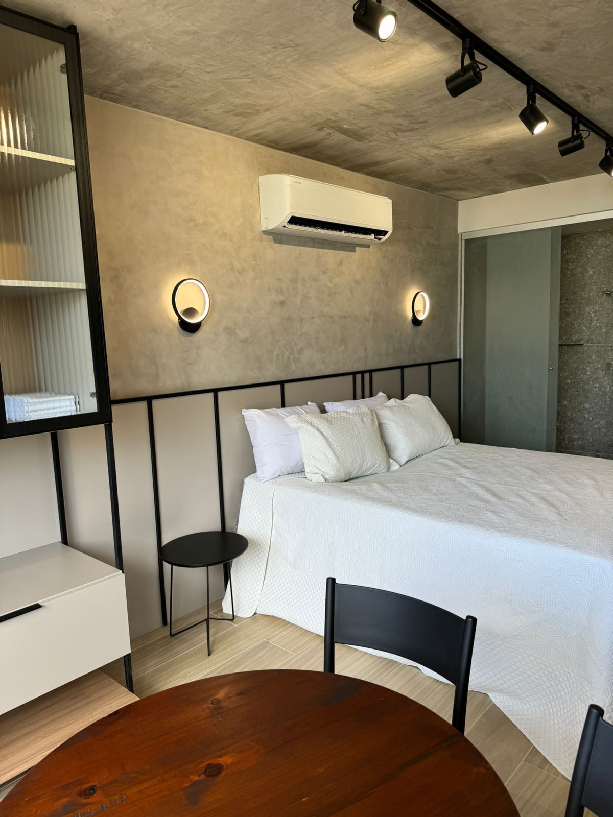 Un dormitorio moderno con cama, mesitas de noche, una mesa redonda de madera y aire acondicionado de pared.
