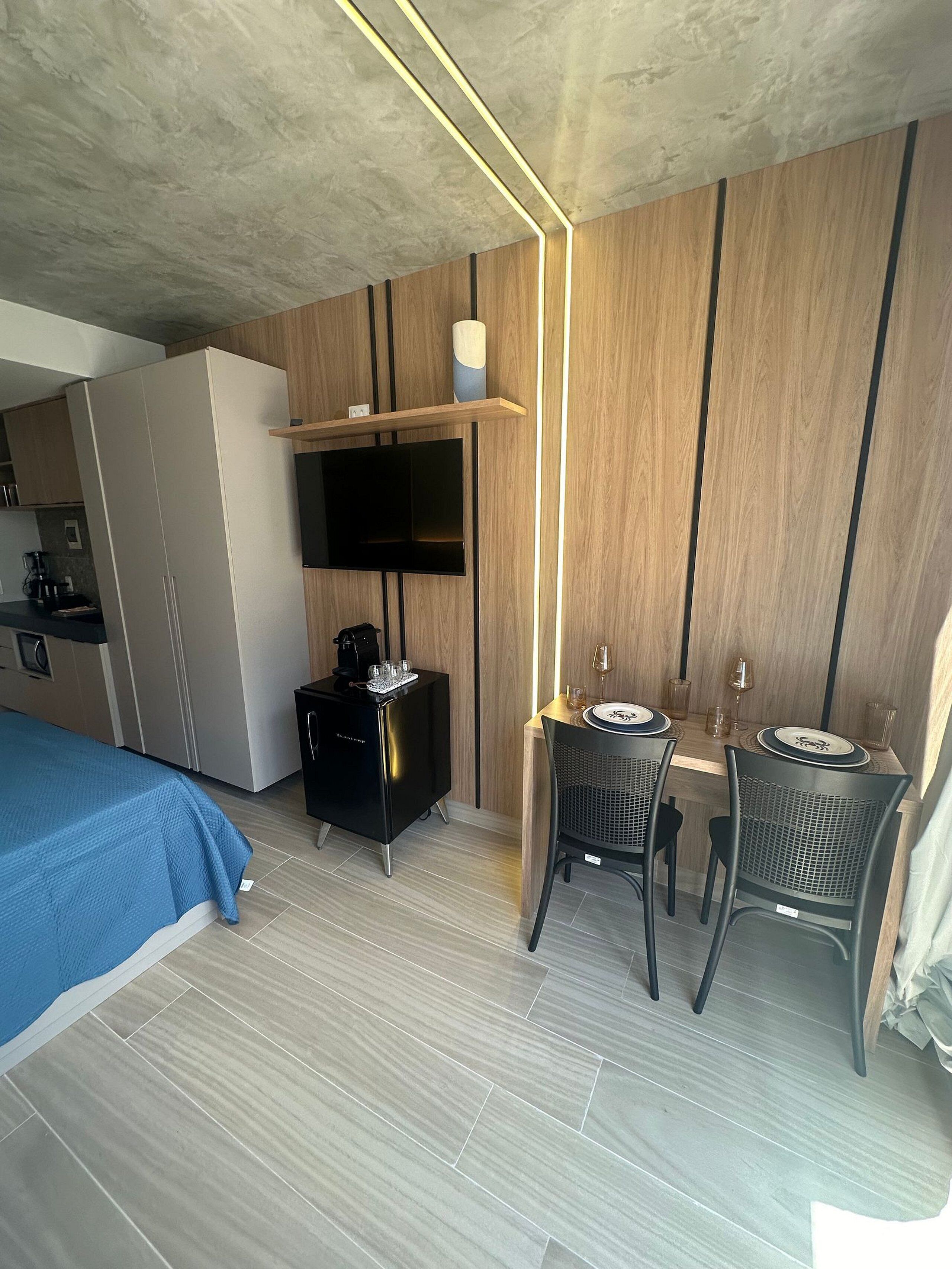 A hotel room with a bed, a desk with two chairs, a TV mounted on the wall, and a coffee machine.