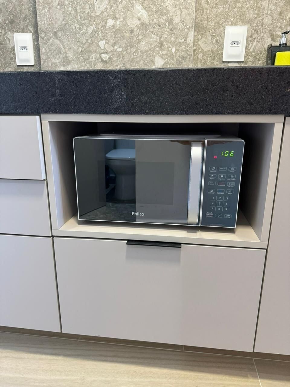 A microwave oven with a digital display, located in a kitchen with a marble backsplash and a black countertop.