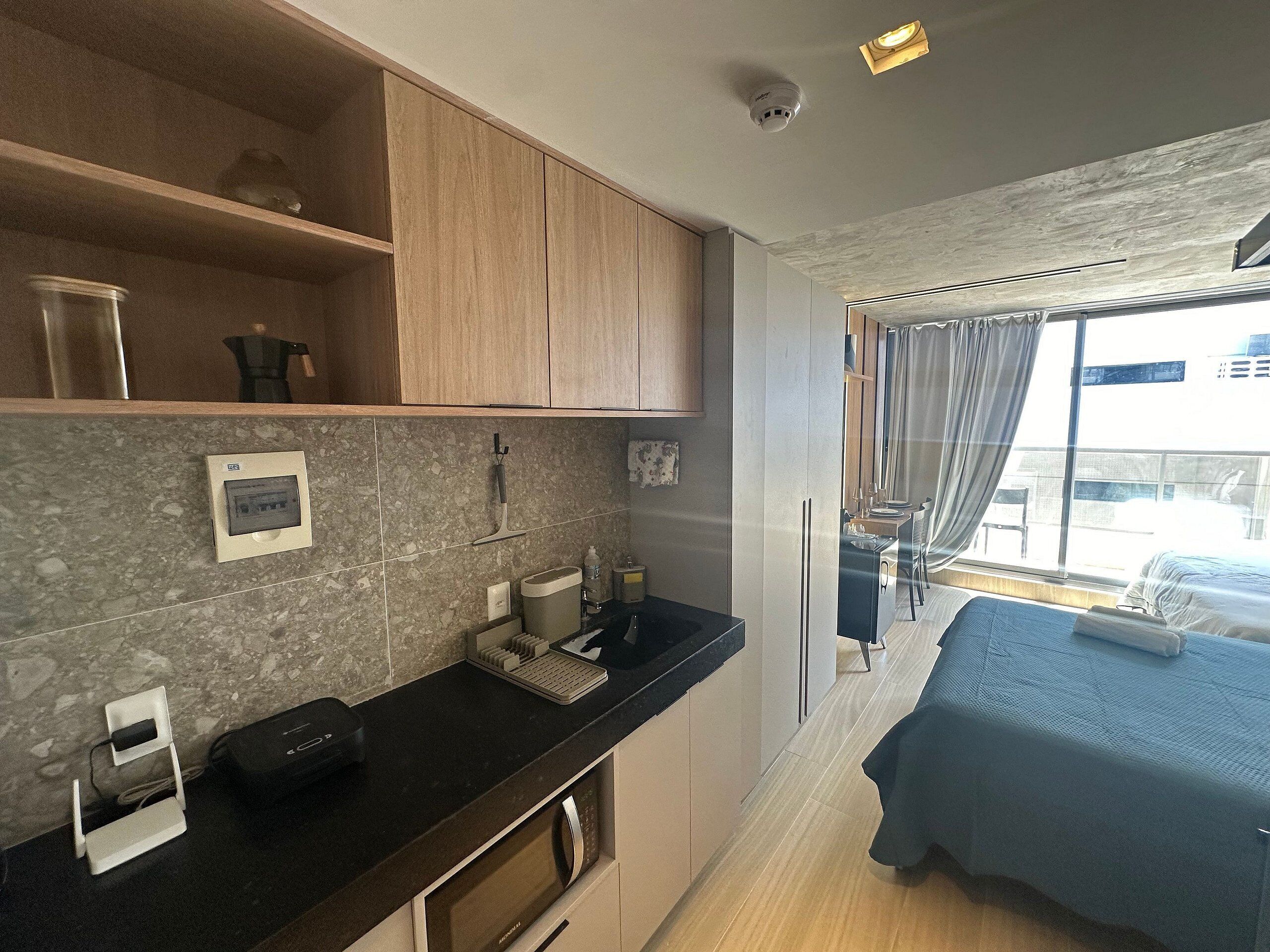 Habitación de hotel pequeña con kitchenette, una cama, ventana con cortinas y vista a la ciudad.