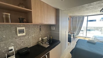 Habitación de hotel pequeña con kitchenette, una cama, ventana con cortinas y vista a la ciudad.