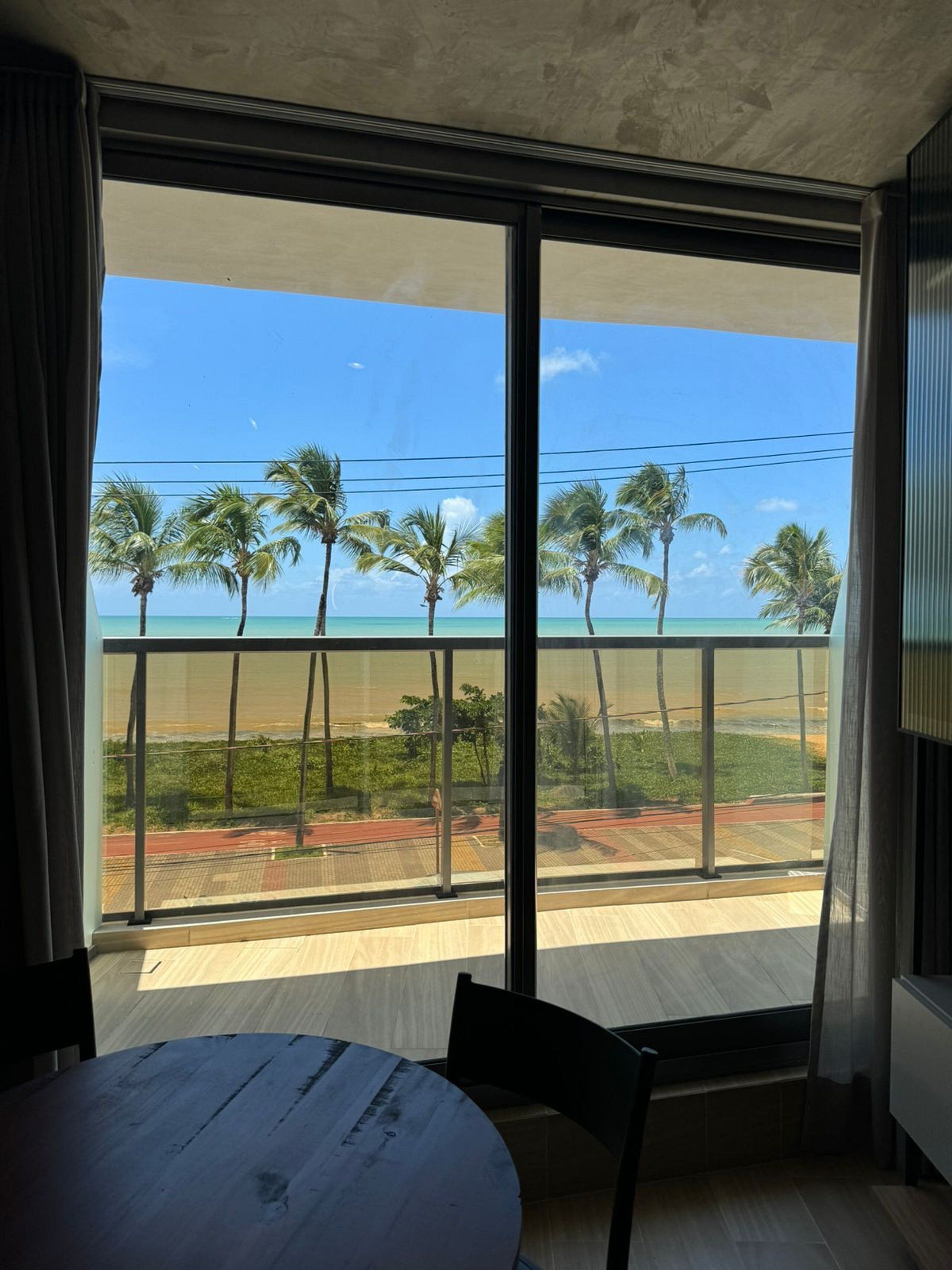 Quarto com uma grande janela com vista para uma varanda com palmeiras e céu claro.