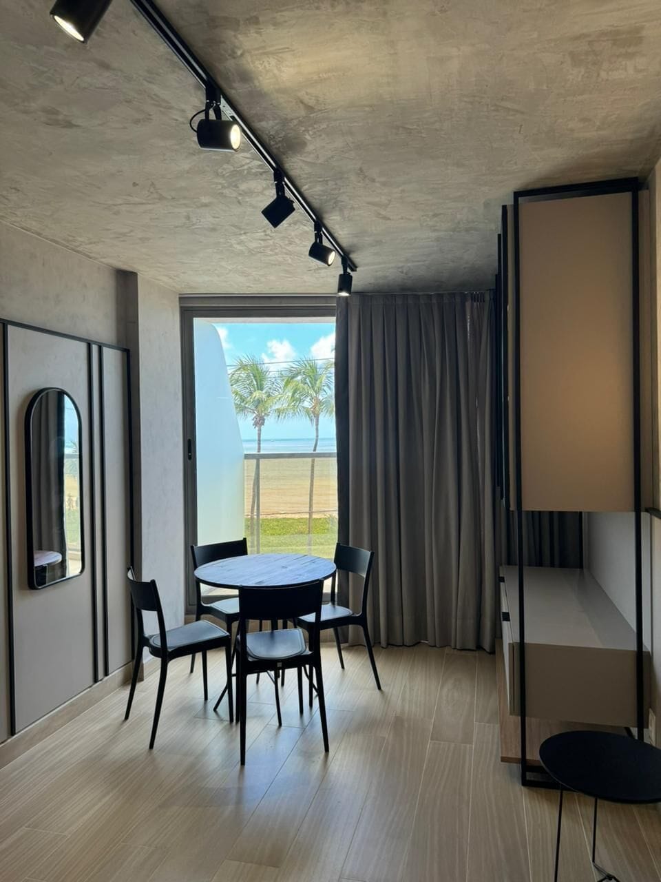 Quarto moderno com mesa redonda, cadeiras, uma janela ampla com vista para a praia e teto com iluminação embutida.