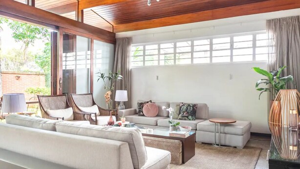 A spacious living room with a sofa, armchairs, a coffee table, and a side table. There is a large window with curtains, a potted plant, and a decorative vase.