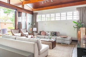 Amplia sala de estar con sofá, sillones, una mesita de centro y una mesita auxiliar. Cuenta con un ventanal con cortinas, una planta en maceta y un jarrón decorativo.