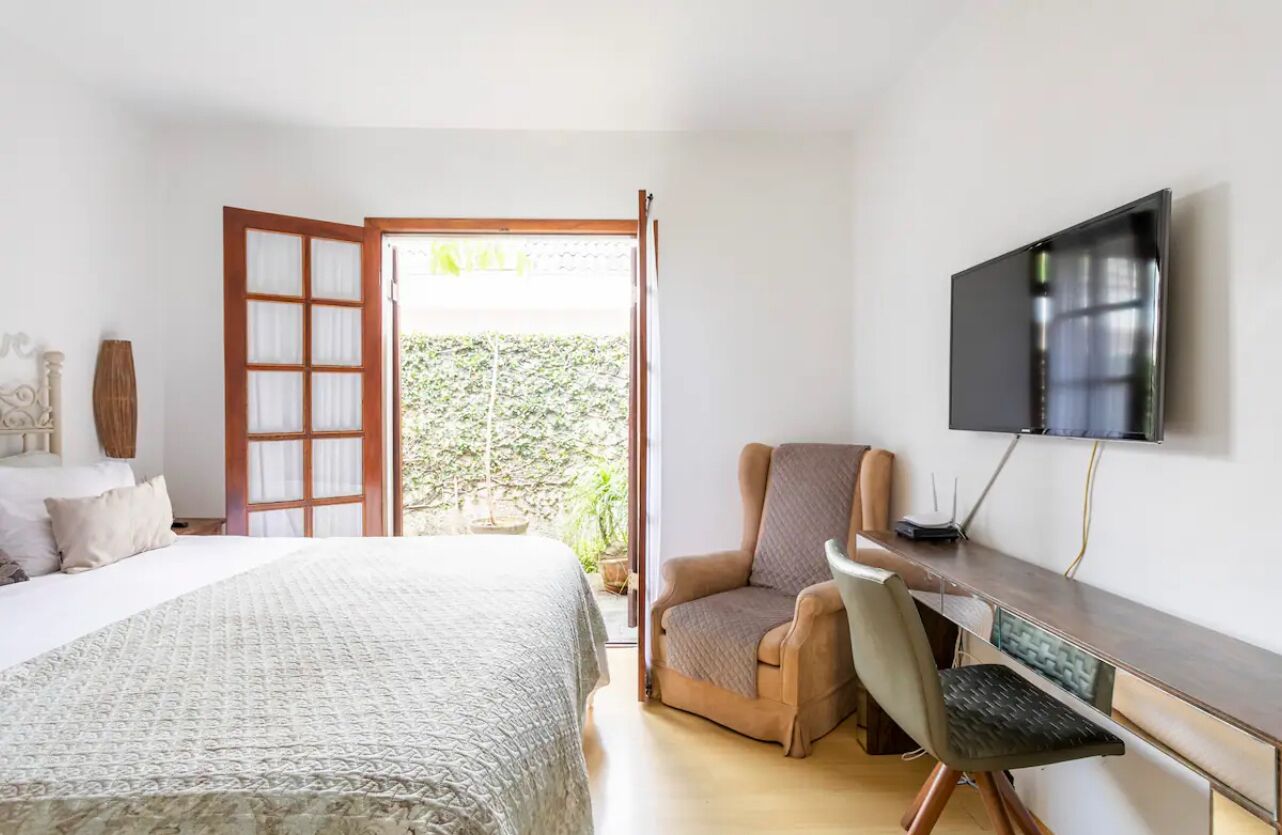 A bedroom with a bed, a desk, a chair, a TV, and a window with a view of greenery.