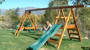 A wooden swing set with a green slide in a grassy area with trees and mountains in the background. - Premium full-hookup drive-through RV 1 space with direct access to  Paiute Trail (Marysvale)