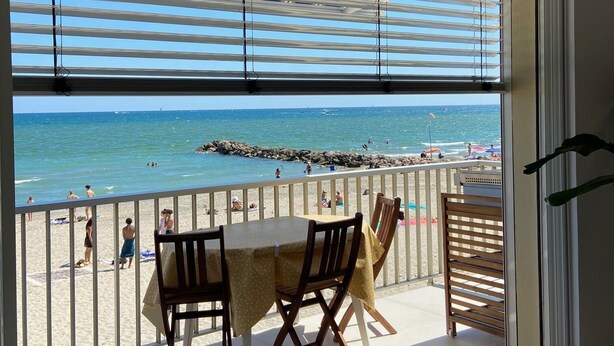 A balcony with a table and chairs overlooking a beach with people and a rocky breakwater. - Appartement Luxe, 1ère Ligne sur la Plage, T3, Climatisé, Terrasse,plancha,wifi (Palavas-les-Flots)