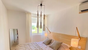 A bedroom with a large bed, a wooden headboard, a bedside table with a lamp, and a window with curtains. - Appartement Luxe, 1ère Ligne sur la Plage, T3, Climatisé, Terrasse,plancha,wifi (Palavas-les-Flots)